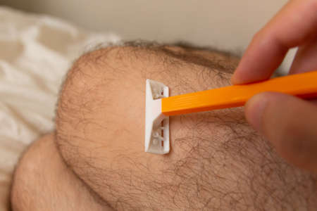 A man removing hair from legs with a razor at home. Man beauty concept. Close up, selective focus and copy spaceの写真素材