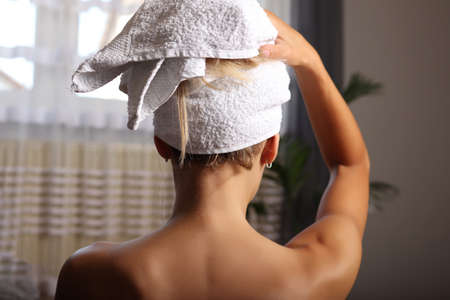 Back view of a woman wrapped in a towel standing next to the window, drying her hair after a morning shower. Hair care conceptの写真素材