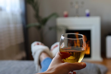 Wine glass in woman hands and she lies in bed and warms legs next to the fireplace on a cold winter dayの写真素材