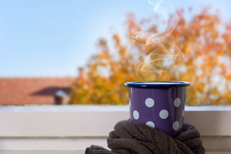 Cup of hot tea wrapped in retro scarf on the window with a blurred autumn view. Close up, selective focus and copy space for design or textの写真素材