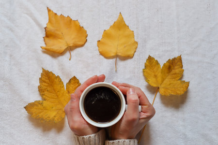 Female hands holding a cup of coffee and fallen yellow leaves on white background. Autumn season...