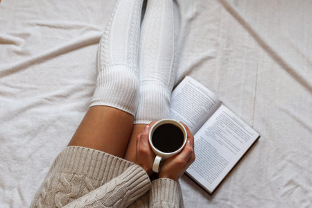 Woman holding a coffee cup on legs in woolen winter socks in a cozy bed with a book at home. Close...
