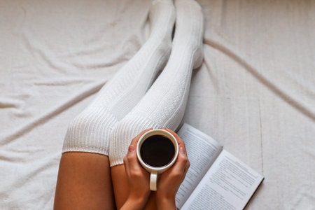 Female hands holding a coffee cup on legs in woolen winter socks in a cozy bed with a book at home. Close up, selective focusの写真素材