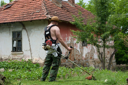Farmer mows tall grass with hands petrol trimmer on a summer day in the garden. A man wears a straw...