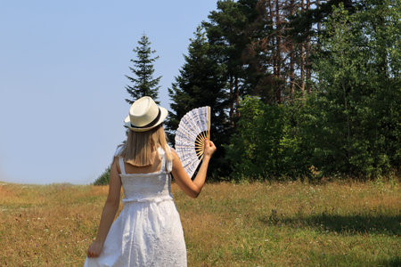 Elegant woman with hand fan cooling and enjoying in beautiful nature with forest and meadow on a hot...