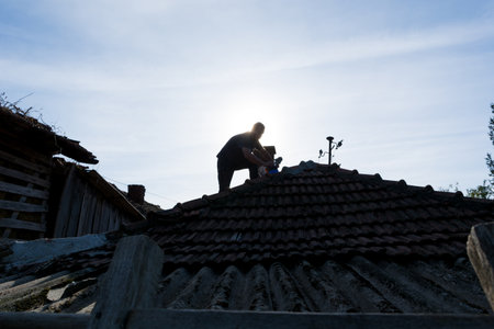 Man on roof fixing tiles with cement on an house roof in the village on a sunny autumn day. Silhouette of a man on a roof while workingの写真素材