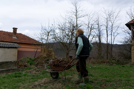 Old woman with a wheelbarrow in front of a rural house