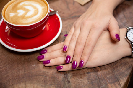 Close-up of female hands with purple manicured nails resting on a wooden table beside a red coffee cup, emphasizing beauty and manicure conceptsの写真素材