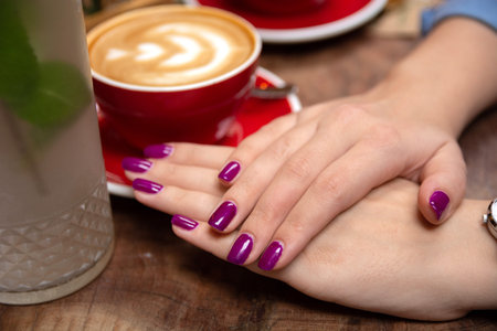 Close-up of female hands with purple nail polish resting on a table, with a background of coffee in a cup. Emphasizes beauty, fashion, and manicure conceptsの写真素材