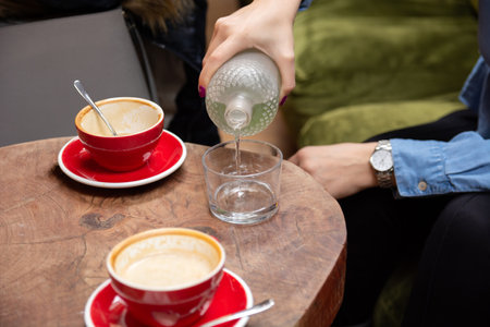 Close-up of a womans hand pouring water from a glass bottle into a glass in a cafe, on a retro wooden table with two red coffee cupsの写真素材