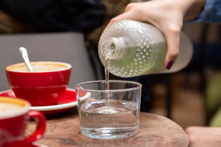 Close-up of a womans hand pouring water into a glass from a glass bottle in a cafe, on a wooden table with coffee cupsの写真素材