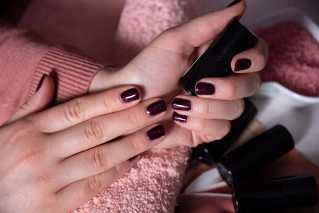 Close-up of female hands with dark purple manicure holding nail polish bottles on a pink towel. Concept of manicure and feminine beautyの写真素材
