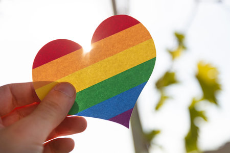 Hand holding a paper heart in LGBT rainbow colors with the sun rays shining around it, concept of...