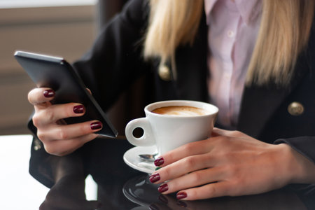 Female hands with a smartphone and coffee cup on a restaurant desk. Woman reading a message on her phone while drinking coffee, close-up, selective focusの写真素材