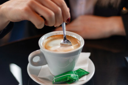 Woman's hand stirring coffee with a small metal spoon on a restaurant desk. Drink concept, close-up, selective focusの写真素材