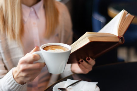 Woman reading a book and drinking coffee at a restaurant table. Wearing a modern pink sweater, concept of relaxation and enjoyment, close-up, selective focusの写真素材