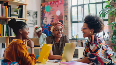 Three young professional women of color are sitting around a table in a brightly lit room. They are all smiling and laughing, and they appear to be enjoying their conversation. The women are all wearing casual clothing, and they are all holding mugs of coffee or tea. The room is decorated with colorful artwork, and there is a large window in the background that lets in natural light. The image is taken from a slightly elevated angle, and it is clear that the women are all friends.の素材