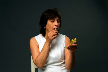 Beauty senior woman in white dress apply lipstick. Studio shot on dark backgroundの写真素材