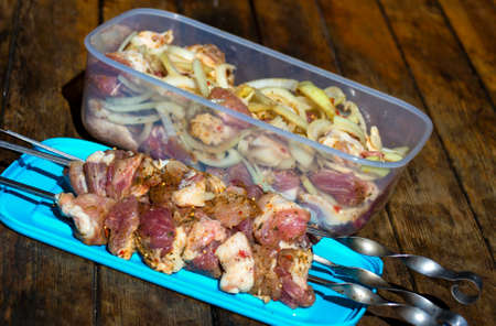 Raw pork meat, making kebab. Skewers ready for grilling and pieces of meat with onion and pepper in plastic food container on wooden table.の写真素材