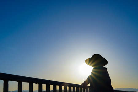 Female Wearing Sun Hat Standing By Railing Taking In The Sunset Silhouetteの写真素材