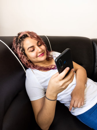 Beautiful girl lying down in an armchair smiling while watching her cell phone.