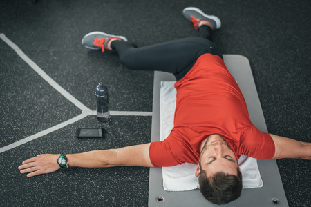 Sporty man stretching back before gym workout. Fitness strong male athlete on floor mat and towel warming up with bottle and cellphone.の写真素材