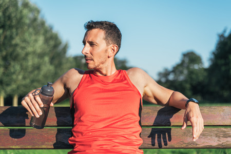 Fitness man resting during outdoor cross training workout with water bottle. Fit fitness sport model sitting on a bench outside using street furniture.の写真素材