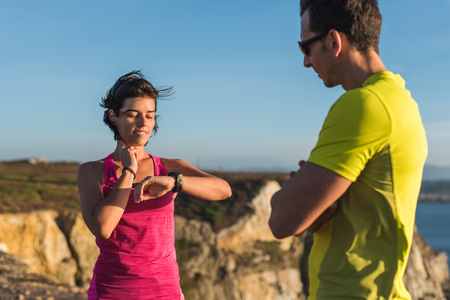 Fitness runner woman checking heart rate pulse with watch and trainer during outdoor trail running workout. Couple team doing intensity workout outside together training for a marathon or decathlon competition.の写真素材