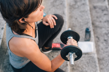 fitness woman lifting weight training arms with dumbbell outside on urban stone stairs with towel, earphones and bottle of water during exercises workout routine. Sporty Female wellness concept.の写真素材