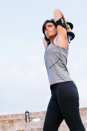 Sport woman lifting weight training arms with dumbbell outside on urban stone stairs with towel, earphones and bottle of water during exercises workout routine. Female wellness concept.の写真素材