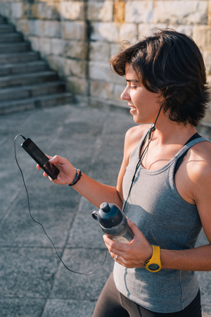 Sportswoman standing resting and listening music on smart phone holding bottle of water while resting from outisde urban training exercise workout. Female wellness concept.の写真素材
