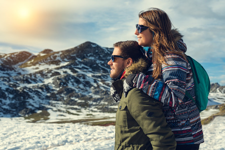 Side view portrait of a pensive couple or marriage hugging and looking to natural snowy landscape wearing gloves, coat and glasses with mountain peak in the background enjoying sun light warmthの写真素材