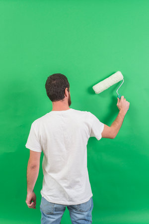 Back view of young man painting wall with paint roller on green backgroundの写真素材
