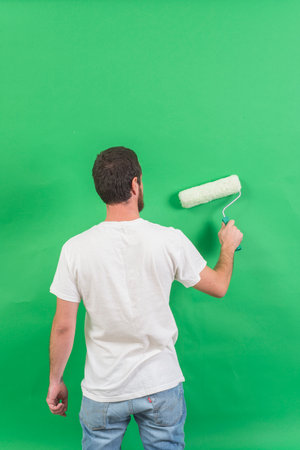 Back view of young man painting wall with paint roller on green backgroundの写真素材
