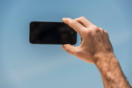 Close-up of male hand taking selfie with smartphone against blue skyの写真素材