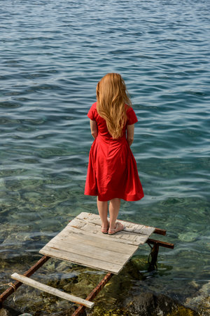 A girl in a red dress stands on a pier in the sea.の写真素材