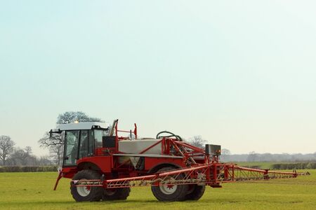 red and white crop spayer in a field of young barley with trees and blue sky backgroundの写真素材