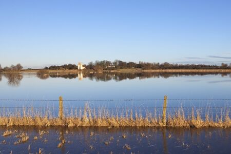 a flooded english landscape with a barbed wire fence and distant ancient church surrounded by trees under a blue skyの写真素材