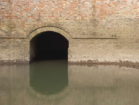 architectural background with an arch tunnel set in an old brick wall which is the outlet from an ancient water mill with a mill pond in the foregroundの写真素材