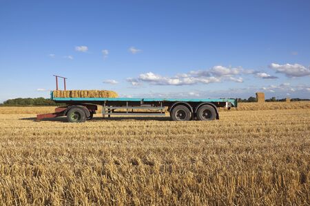 agricultural landscape with long straw bale trailer parked in a stubble field with haystack and trees under a blue summer sky with white fluffy cloudsの写真素材