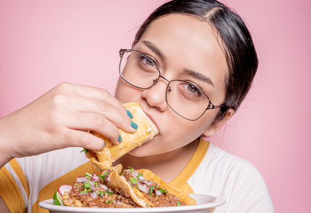 young mexican woman eating mexican style birria tacos, with a yellow shirt and a bright pink backgroundの写真素材