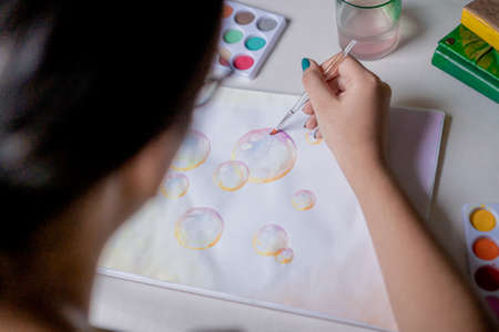 Woman painting colorful bubble drawing in a sketchbook with colored watercolors on a white table.の写真素材
