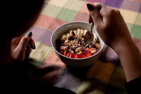 Woman eating delicious homemade oatmeal with banana, strawberries, granola, blueberry, coconut and almonds with a spoon inside a white plate on a table.の写真素材
