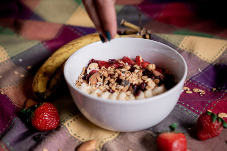 Delicious homemade oatmeal with banana, strawberries with one hand pouring granola, blueberry,...