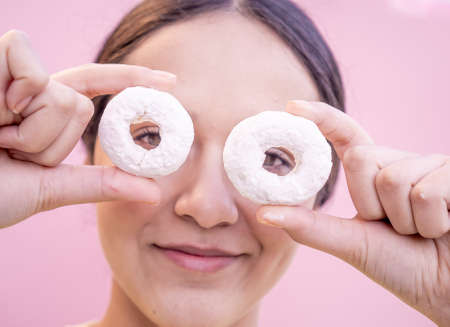 Latin woman holding two delicious donuts sprinkled with sugar glass in front of her eyes with a pink background in close up concept.の写真素材