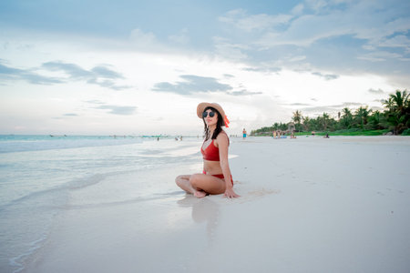 Beautiful Latina woman in an orange swimsuit and white hat sitting on the beach looking out to sea...