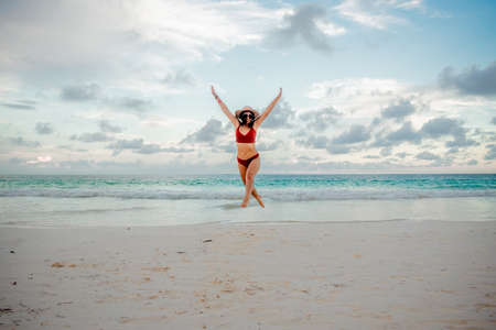 Beautiful Latina woman in an orange swimsuit and white hat on the beach with her hands raised and the sea behind her in Quintana Roo.の写真素材