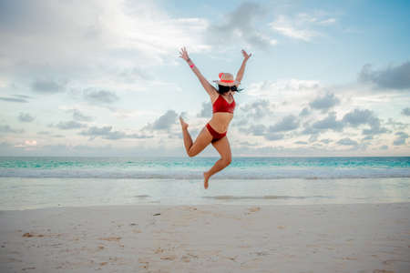 Beautiful Latina woman in an orange swimsuit and white hat jumping on the beach with the sea behind her in Quintana Roo Mexico.の写真素材