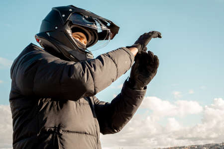 Motorcyclist wearing a black dual-purpose motorcycle helmet with a black jacket with a sky background wearing motorcycle gloves.の写真素材