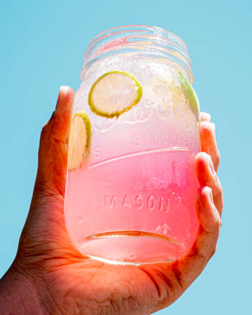 Refreshing pink lemonade in a glass tumbler with ice and lemon slices, held in one hand, with the sky in the background.の写真素材